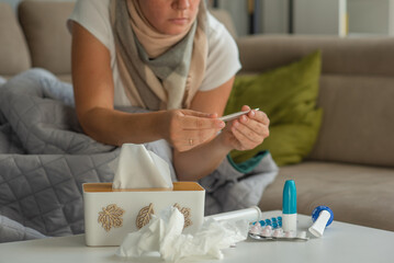 Medicines against colds and paper napkins are on the table, a woman with signs of flu and colds is sitting in the background and holding a thermometer in her hands.