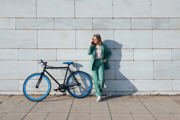 Young empowered ginger-haired woman in green suit talking on smartphone standing with bicycle at...