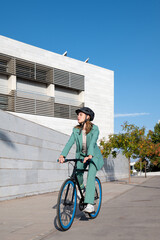 Stylish business woman in green suit and helmet riding her bicycle at the financial district with modern building on the background