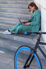 Business woman sitting on stairs outside office building while working with laptop next to a bike