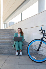 Business woman sitting on stairs outside office building while working with laptop next to a bike