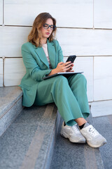 Business woman sitting on stairs outside office building while using her smartphone