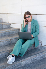 Business woman sitting on stairs outside office building while working with laptop and having a phone call