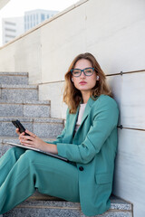 Business woman sitting on stairs outside office building looking at camera while using her earphones