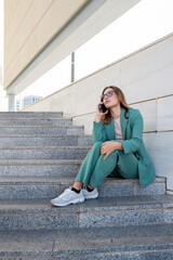 Business woman sitting on stairs outside office building while working with laptop and having a phone call