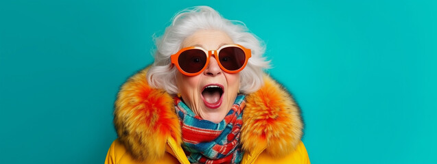 Colorful studio portrait of eccentric elderly granny wearing orange fur and sunglasses, blue background - obrazy, fototapety, plakaty