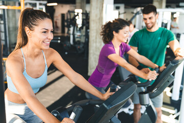 Fototapeta na wymiar Young smiling woman with ponytail wearing fitness clothes riding a gym bike while being accompanied by her less experienced female friend.