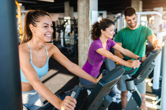 Smiling Caucasian female athlete practicing on exercise bike in the gym. Two attractive girls doing cardio under the advisory of their fitness instructor.