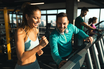 Fototapeta na wymiar Young athlete male with headphones assisting a young woman while exercising on treadmills in a gym. Two people having engaging conversation during workout.