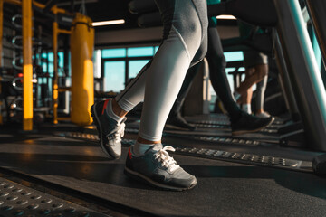 Unrecognizable athletes running on treadmills in health club. Bellow knees view of female legs captured in motion while doing cardio. - obrazy, fototapety, plakaty