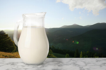 Fresh milk in glass pitcher on white marble table against mountain landscape, space for text