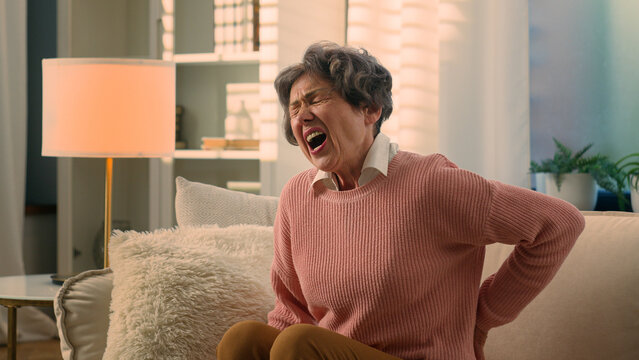 Caucasian old woman senior lady mature retirement granny elderly female at couch home try to stand up feel painful spasm backache suffer with pain in back spinal ache injury spine osteoporosis sore