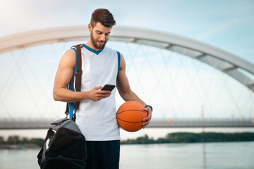 Young tall sportsman walking with sports bag while using mobile phone and holding a basketball....