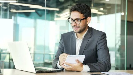 Young businessman in a formal suit listens to online e-learning, video course sitting in office. Man at a distance learning seminar writes in a notebook using a laptop computer communicates with coach