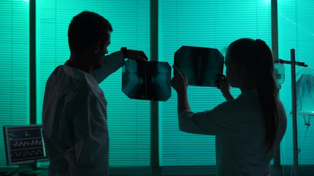 Medium-sized silhouette video of two doctors standing in a hospital ward. Doctors look at patient's leg, knee x-ray scan, discussing the diagnosis and a treatment method.