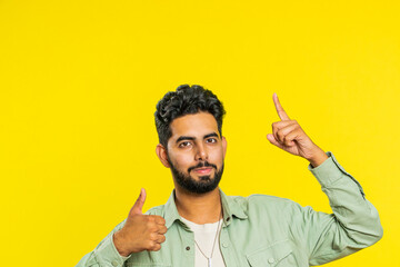 Indian bearded man showing thumbs up and pointing overhead, above head empty place, advertising area for commercial text copy space for goods promotion advertisement. Guy isolated on yellow background