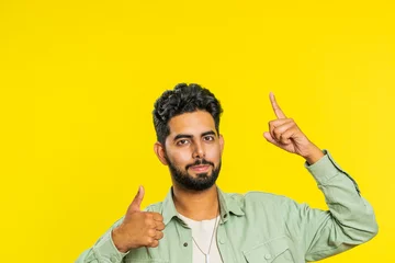 Fotobehang Indian bearded man showing thumbs up and pointing overhead, above head empty place, advertising area for commercial text copy space for goods promotion advertisement. Guy isolated on yellow background © Andrii Iemelianenko