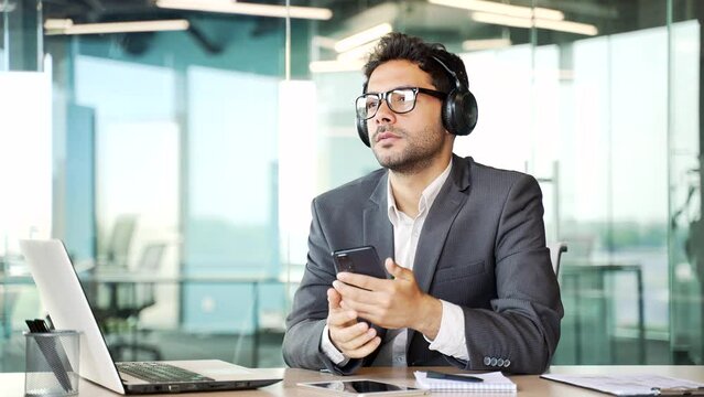Young businessman with headphones listening to relaxing music sitting in a business office. Happy entrepreneur turns on a playlist on smartphone and enjoys the sounds of relaxation and noise of nature