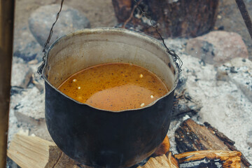 Metal cauldron with mouthwatering soup hanging on tripod under firewood before cooking on fire in...