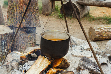 Metal cauldron with meal inside hanging on tripod over firewood, preparing for boiling on bonfire...