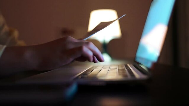 Close up video of Caucasian woman in casual yellow shirt checking an electricity bill, she is making a contactless payment using her smartphone, sitting at table with laptop computer at night