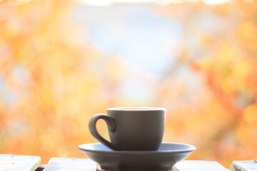 A cup of Americano coffee on a wooden white table on a windowsill in autumn with a maple tree of colorful leaves in the garden with copy space. Seasonal autumn and beautiful relax concept