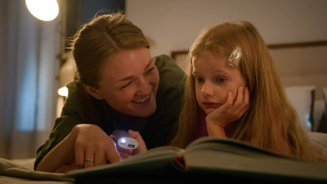 Caring caucasian mother reading book with little kid daughter happy family resting on bed in bedroom at home mom and child girl enjoy bedtime fairytale story read stories in evening with flashlight