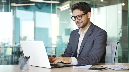 Young businessman in a formal suit typing on a laptop while sitting at a workplace in business office. Handsome entrepreneur working on a project on the computer, texting a client or chatting online - Powered by Adobe