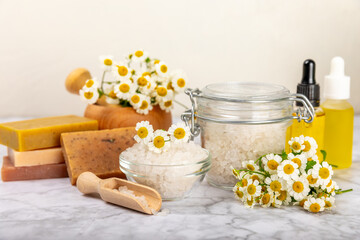 Chamomile spa. Composition with chamomile flowers, handmade soap, essential oil cosmetic bottle, body cream, scrub and sea salt on a white texture background. Relaxing beauty treatments.Copy space