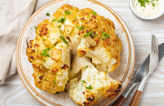 Whole Roasted Head Of Cauliflower With Olive Oil On Plate On White Wooden Rustic Table Top View With Cutlery. Vegetarian Dish..