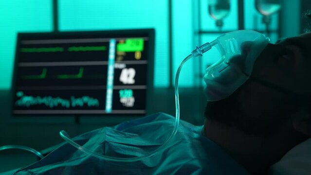 Close-up detail video of a patient on breathing support dying in a hospital bed. His ICU monitor on the background displays loss of heart and brain activity.