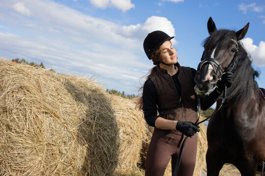 Equestrian sports. A young woman in a sports uniform, a rider and her horse next to a stable and haystacks