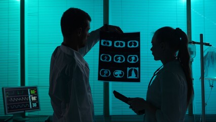 Medium-sized silhouette shot of two doctors standing in a hospital ward. Doctors look at patient's...