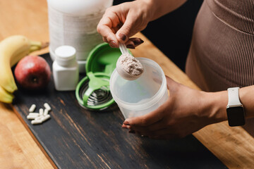 Athletic woman in sportswear with measuring spoon in her hand puts portion of whey protein powder into a shaker on wooden table with amino acid white capsules, bananas and apple, making protein drink