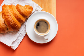 White cup of hot aromatic coffee and fresh french croissant on a wooden desk from above on an orange background. Breakfast idea.