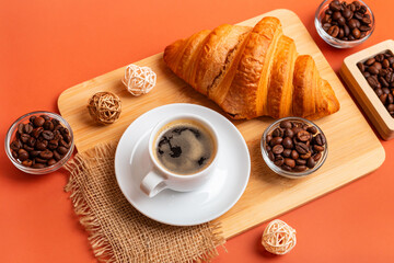 French croissant, cup of black coffee, aromatic roasted arabica and robusta coffee beans in wooden box and glass jars on  wooden desk from above on orange background in rustic style. Perfect breakfast