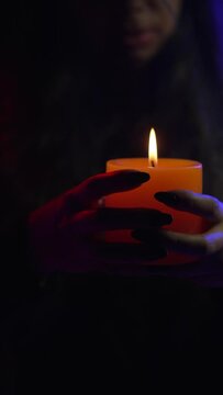 Woman dressed in black lace in a dark environment, holds an orange candle. Her nails painted black. Close up shot. Halloween concept. Vertical