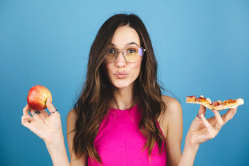 Confused young brunette curly woman choosing slice of pizza instead of red apple. Close up portrait...
