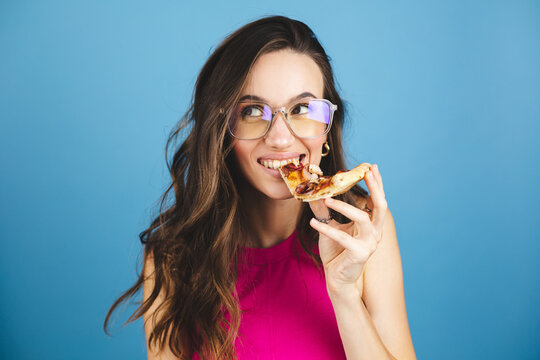 Woman eats slice of pizza, dressed in pink top, isolated blue background. Pretty girl has snack with fast food. Pizza and delivery concept. Happy woman posing with pizza slice, look happy, bite pizza.