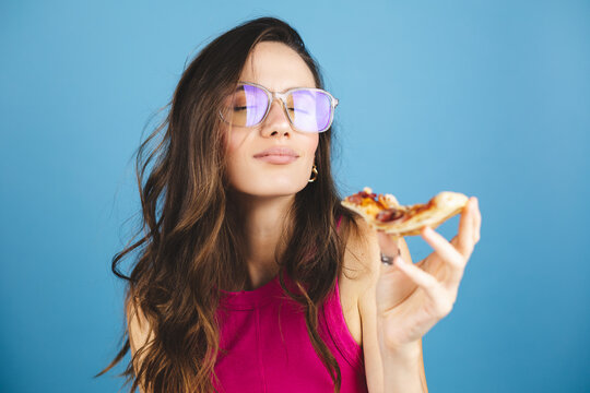 Brunette curly woman enjoys delicious slice of pizza, likes this taste, closes eyes from pleasure, has good appetite, dressed in pink top, isolated over blue background. Hungry woman indoor.