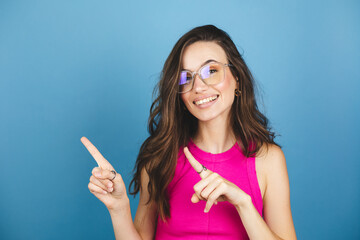 Look, advertise here! Dark haired pretty woman with appealing smile gives positive attitude towards good deal, expresses her recommendation while pointing on left side isolated on blue background.