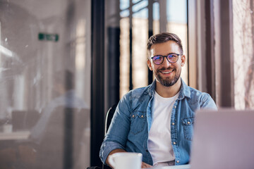 Handsome smiling young man drinking coffee sitting at office desk and looking at camera