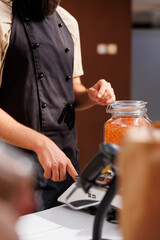 Storekeeper at checkout counter in zero waste shop weighting vegan products purchased by shopper. Client purchasing healthy food in local neighborhood store with paper bags only policy, close up