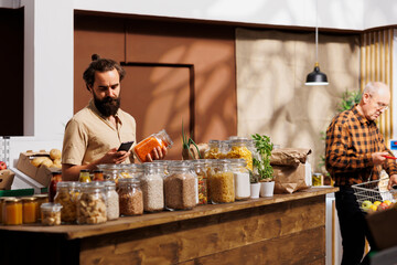 Man in zero waste store analyzing bulk products, using phone to make sure they are freshly harvested. Sustainable living customer thoroughly checking local supermarket food items are additives free