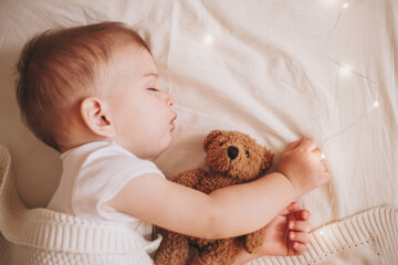 A little baby in a white bodysuit sleeping in a white crib holding a teddy bear and surrounded by small lights