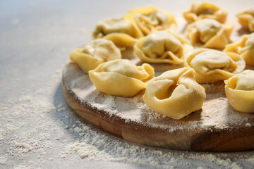 Raw traditional ravioli tortellini on wooden plate, grey table with flour