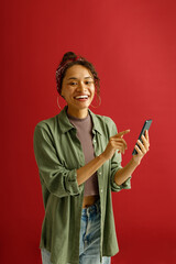 Smiling woman is pointing on mobile phone while standing on red studio background