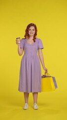 A redhaired woman holds shopping bags and a paper cup of tea or coffee. Young woman in full length sundress isolated on yellow. Vertical shot. Concept of selling discounts on purchases.