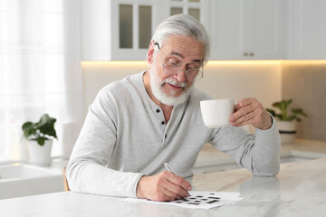 Senior man with cup of drink solving crossword at table in kitchen
