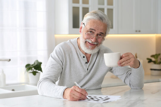 Senior man with cup of drink solving crossword at table in kitchen